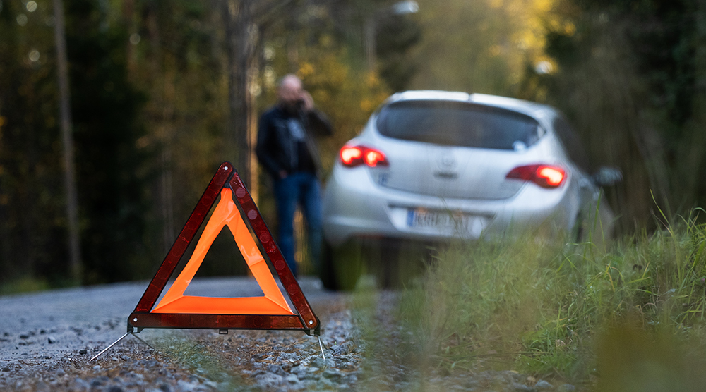 Kuvituskuva, jossa tien varteen pysähtyneen auton takana on varoituskolmio, ja henkilö puhuu matkapuhelimeen.