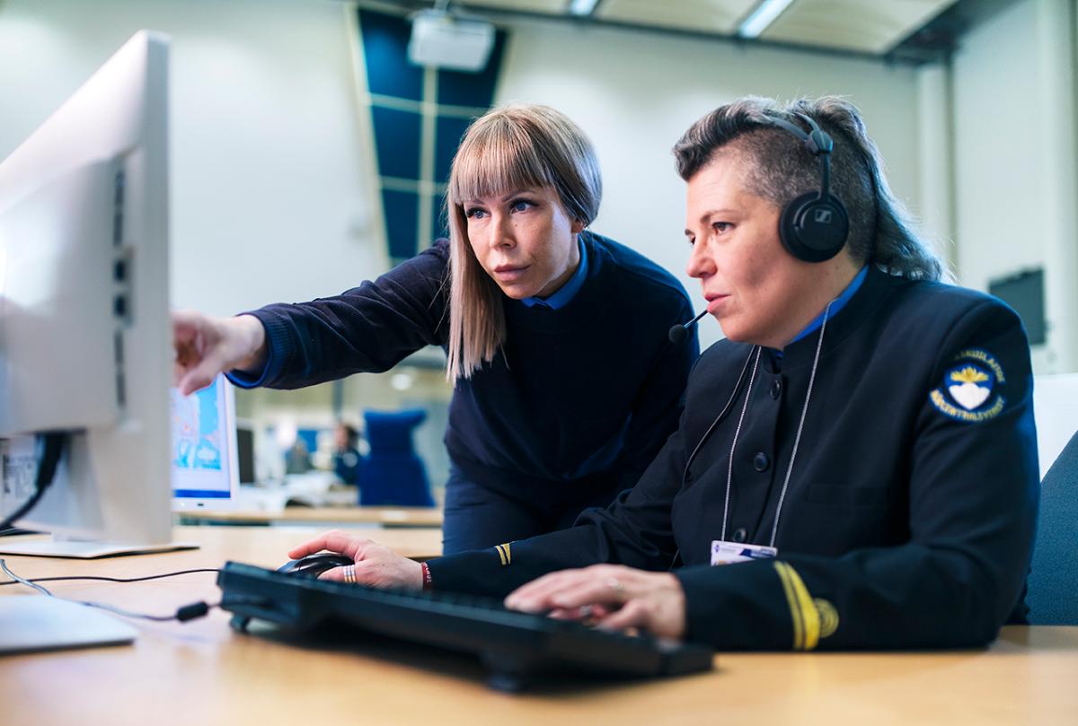 Två kvinnor i uniformer som nödcentraloperatör framför datorskärmar. En av dem har ett headset på sig och den andra står och pekar med ett finger mot skärmen.