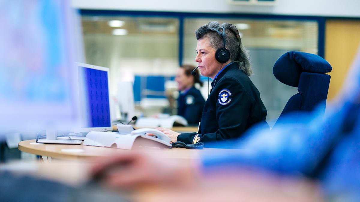 Nainen Hätäkeskuspäivystäjän vEn kvinna i uniform av en nödcentralsoperatör framför en skärm med en headset-mikrofon på huvudet.