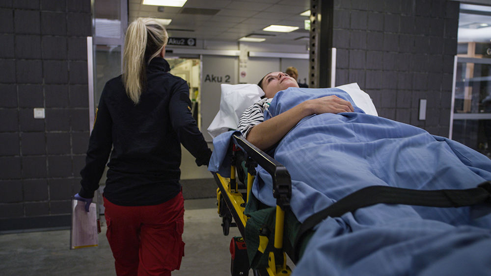 Kuvassa ensihoitaja ohjaa paareja kohti sairaalan ovia. Paareilla makaa potilas.