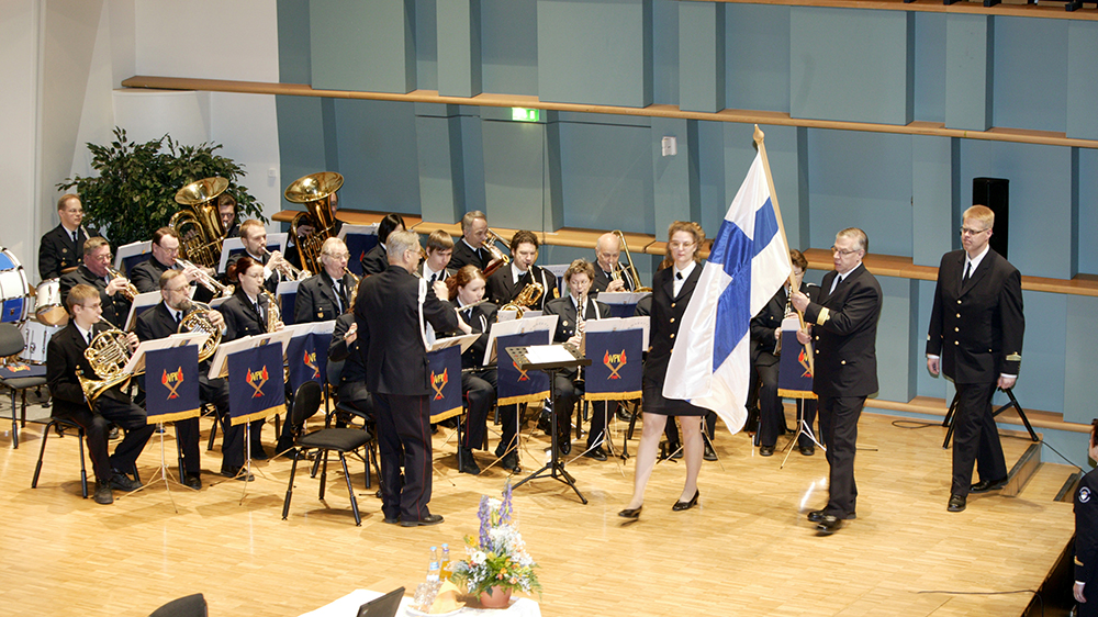 Lavalla kävelevä virkamies kantaa Suomen lippua ja taustalla on orkesteri.