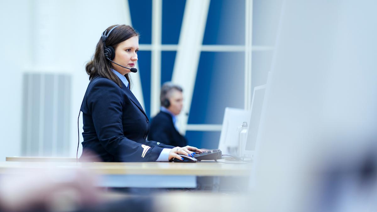 En kvinna sitter framför en dator och har hörlurar på. Kvinnan bär Nödcentralsverkets uniform. En annan person visas i bakgrunden.