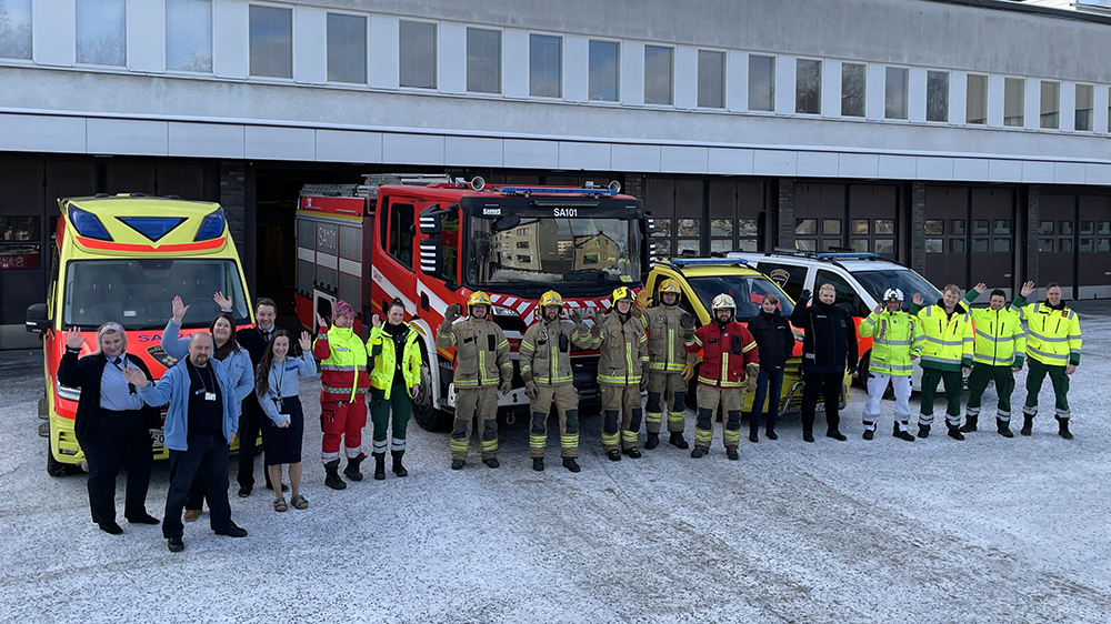 Hätäkeskuspäivystäjiä sekä pelastus- ja ensihoitohenkilöstöä yhteiskuvassa. Taustalla hälytysajoneuvoja.