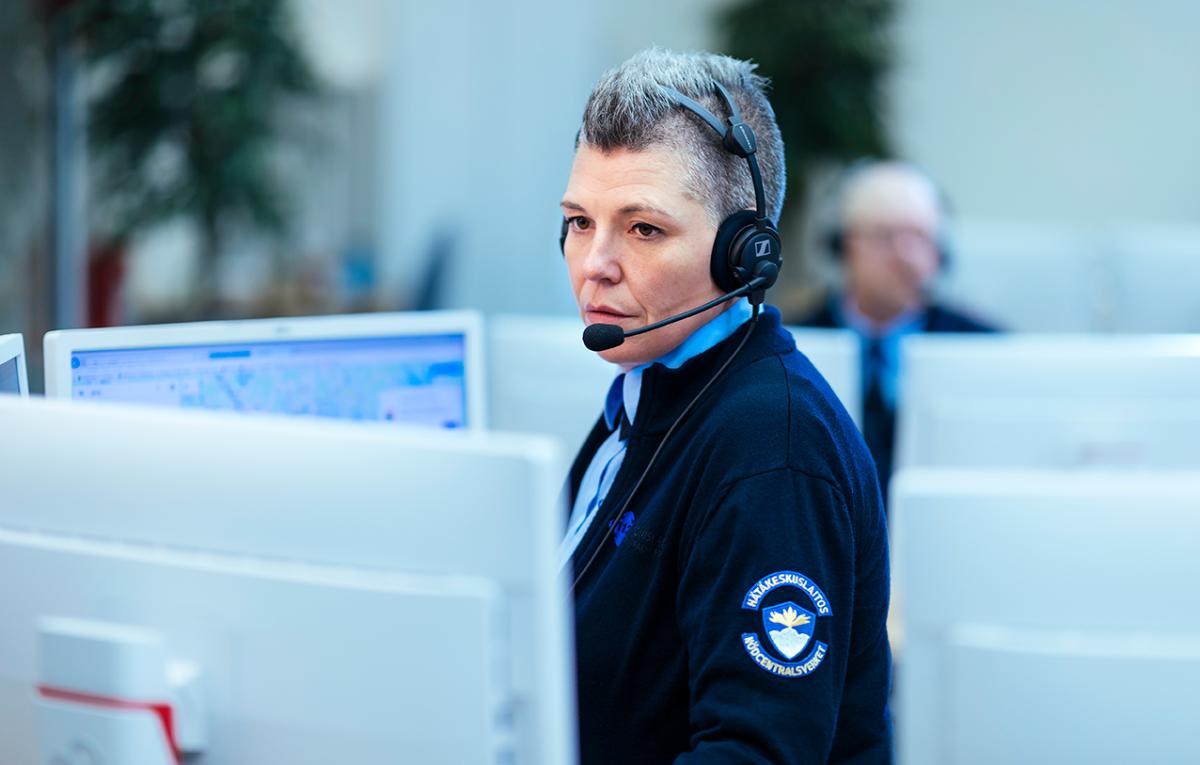 En nödcentralsoperatör på jobbet framför datorskärmar med hörlurar på öronen som tar emot ett nödsamtal.