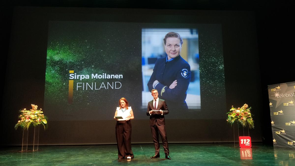 Sirpa Moilanen håller festtal med nödnummerorganisationens representant bredvid sig. I bakgrunden syns en bild på Sirpa iklädd arbetskläder.