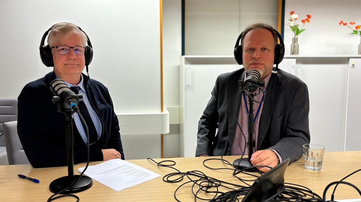 Ari Ekstrand ja Lasse Ilkka istuvat vierekkäin pöydän ääressä ja hymyilevät kohti kameraa. Molemmillaan on edessään mikrofoni ja päässään kuulokkeet.