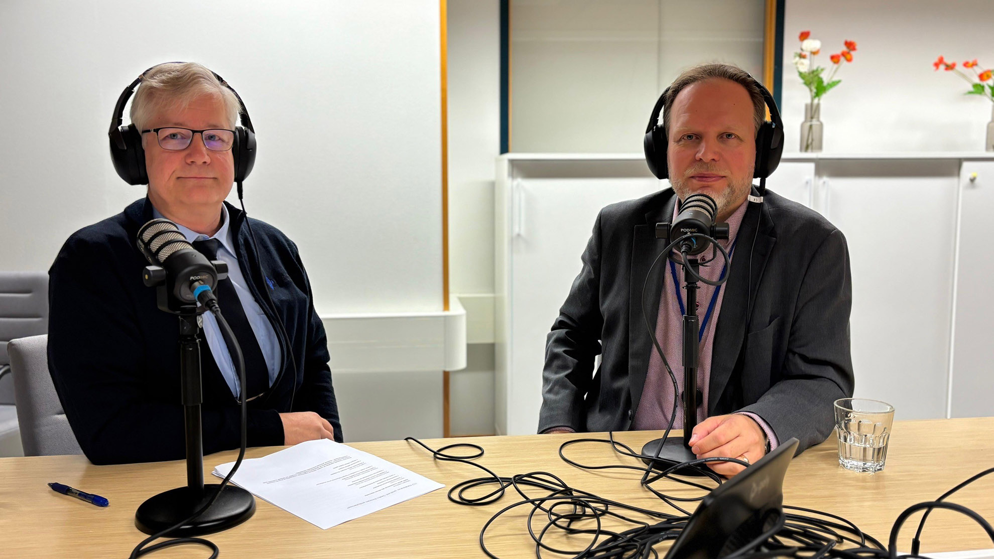 Podcastin vieraat istuvat vierekkäin ja hymyilevät. Heillä on kuulokkeet päässä ja mikit kasvojen edessä. Pöydällä kiemurtelee studion johtoja.