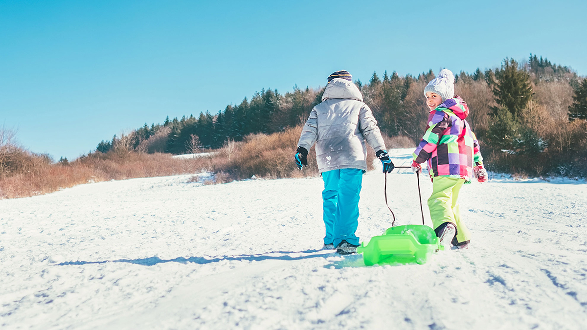 Kaksi lasta vetää pulkkaa lumisessa maisemassa.