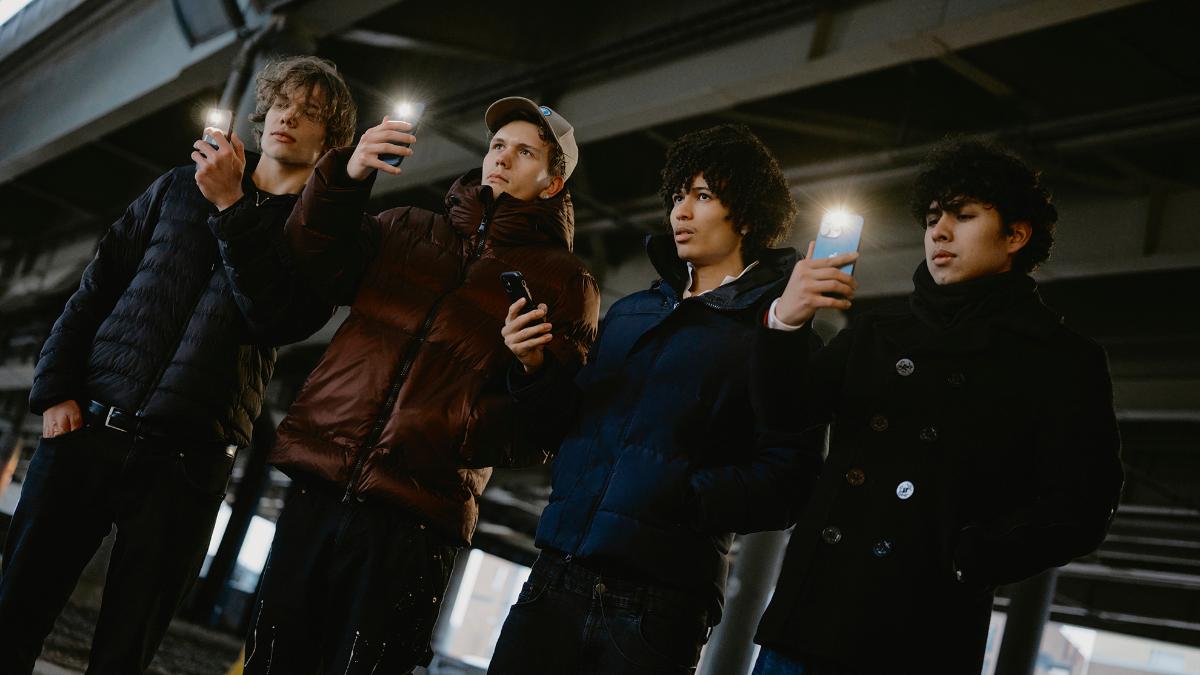 Neljä nuorta miestä seisoo vierekkäin ulkona. Kolme heistä kuvaa puhelimella, mutta yksi katsoo mietteliäänä eteenpäin puhelin kädessään.