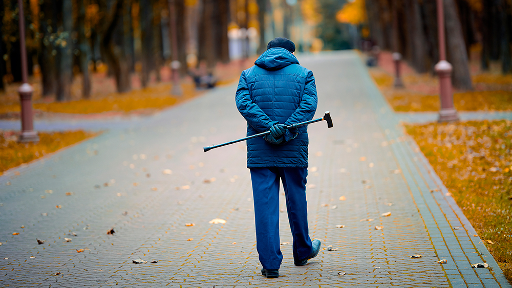 Vanha mies kävelee kädet selän takana kävelykeppiä pidellen syksyisessä puistossa.
